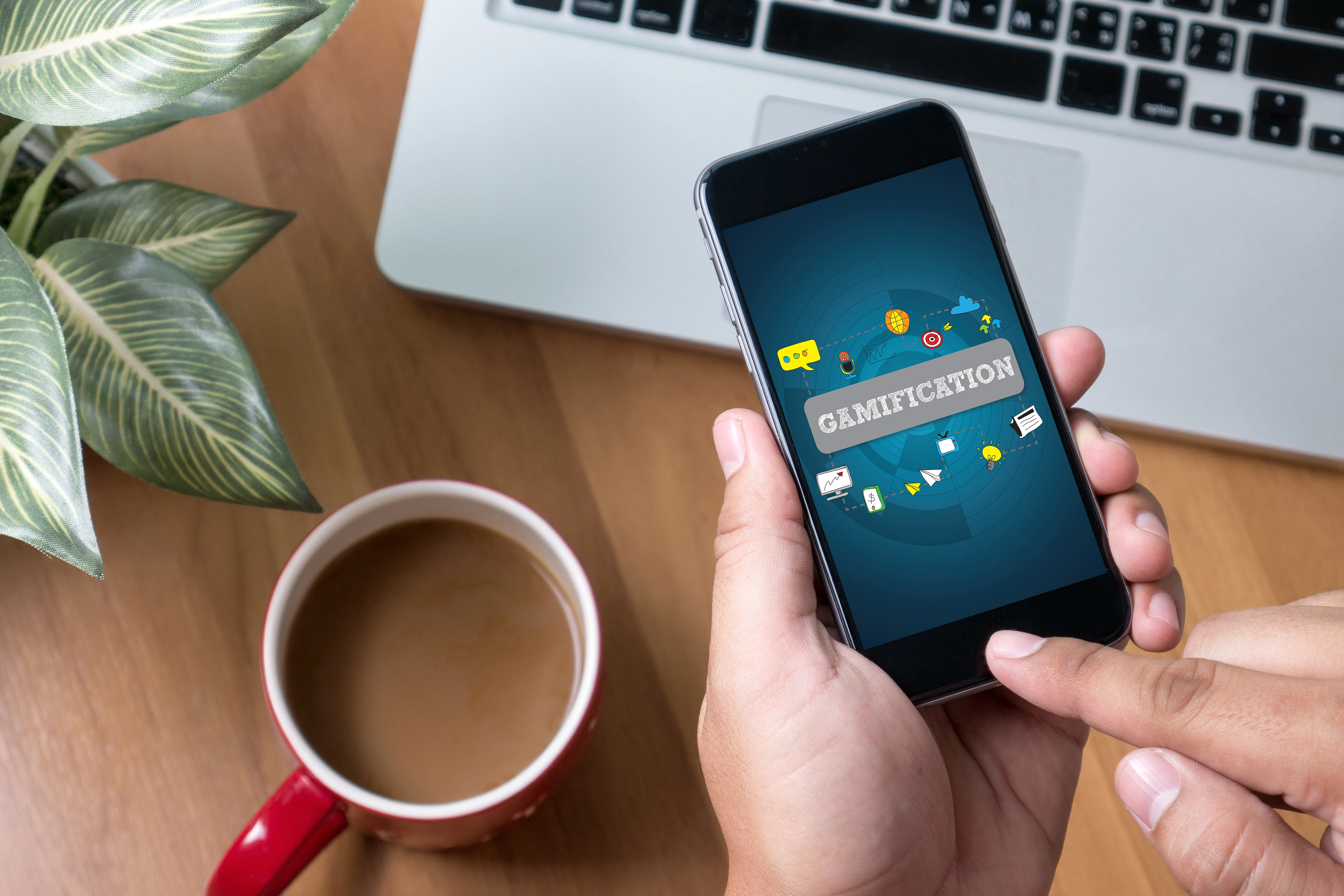Person sitting with a laptop and cup of coffee showing gamification on their phone